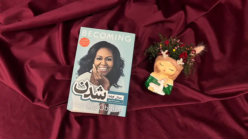 کتاب خاطرات و زندگینامه میشل اوباما روی یک میز در کنار یه گلدان با گل جاودان